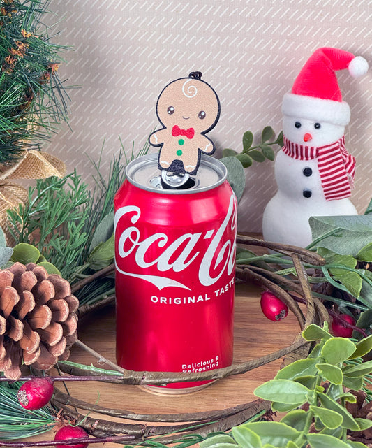 Coca-Cola can with a gingerbread man Tab Buddy on top, surrounded by Christmas decorations.