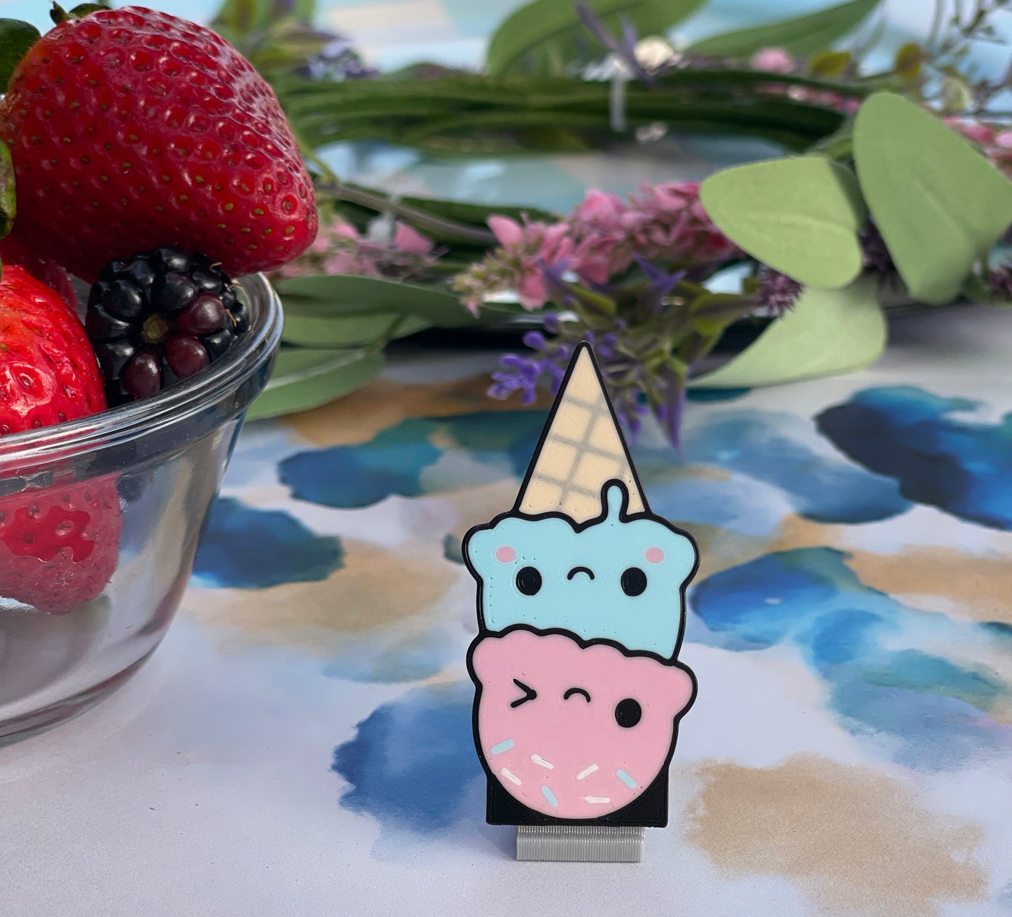 Kawaii-style ice cream cone Tab Buddy with a scoop of pink and scoop of blue ice cream with smiley faces on a beige ice cream cone on a colorful surface with strawberries and flowers.
