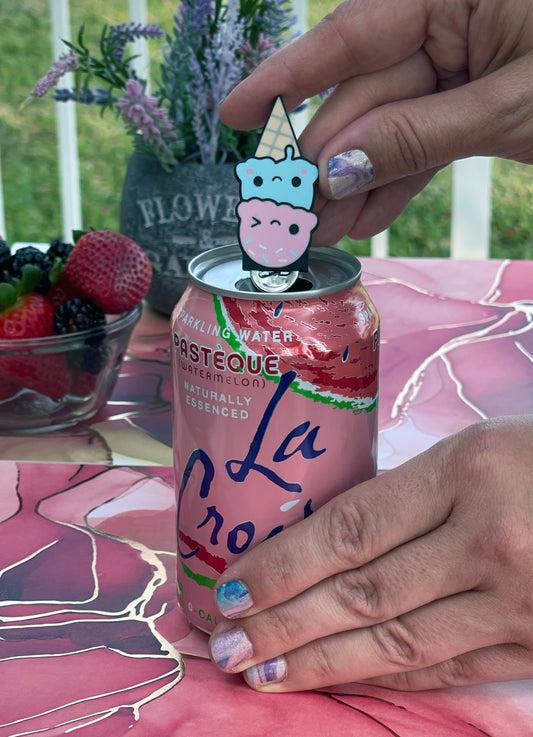 Person holding a pink soda can with a ice cream Tab Buddy on top, surrounded by strawberries and flowers on a pink surface.