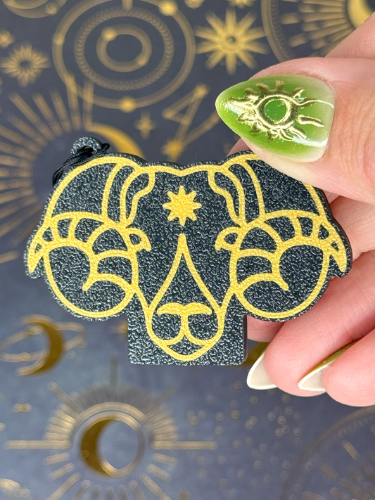 Decorative black and gold Aries Tab Buddy held by a hand with green nail polish against an astrological background.