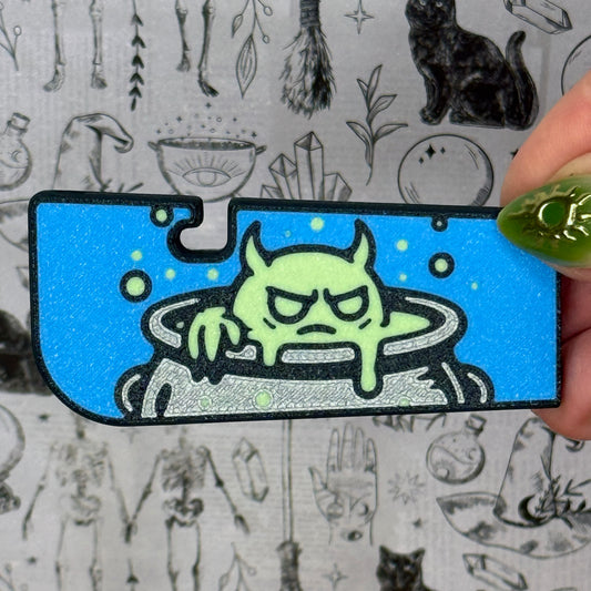 Tab Buddy XL with a green creature in a cauldron design on a blue background, held by a hand against a black and white patterned background.