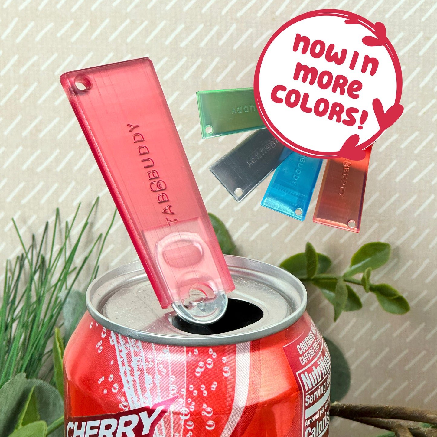 Ruby red translucent Tab Buddy Classic sits atop a cherry soda can, surrounded by greenery.