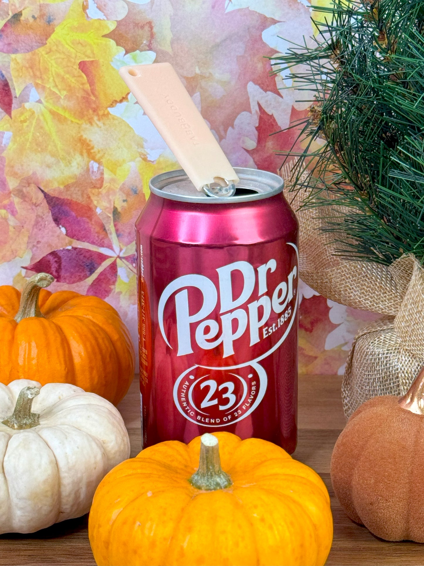 A peach-colored Tab Buddy Classic sits atop a Dr Pepper can with pumpkins and decorative elements on a colorful background