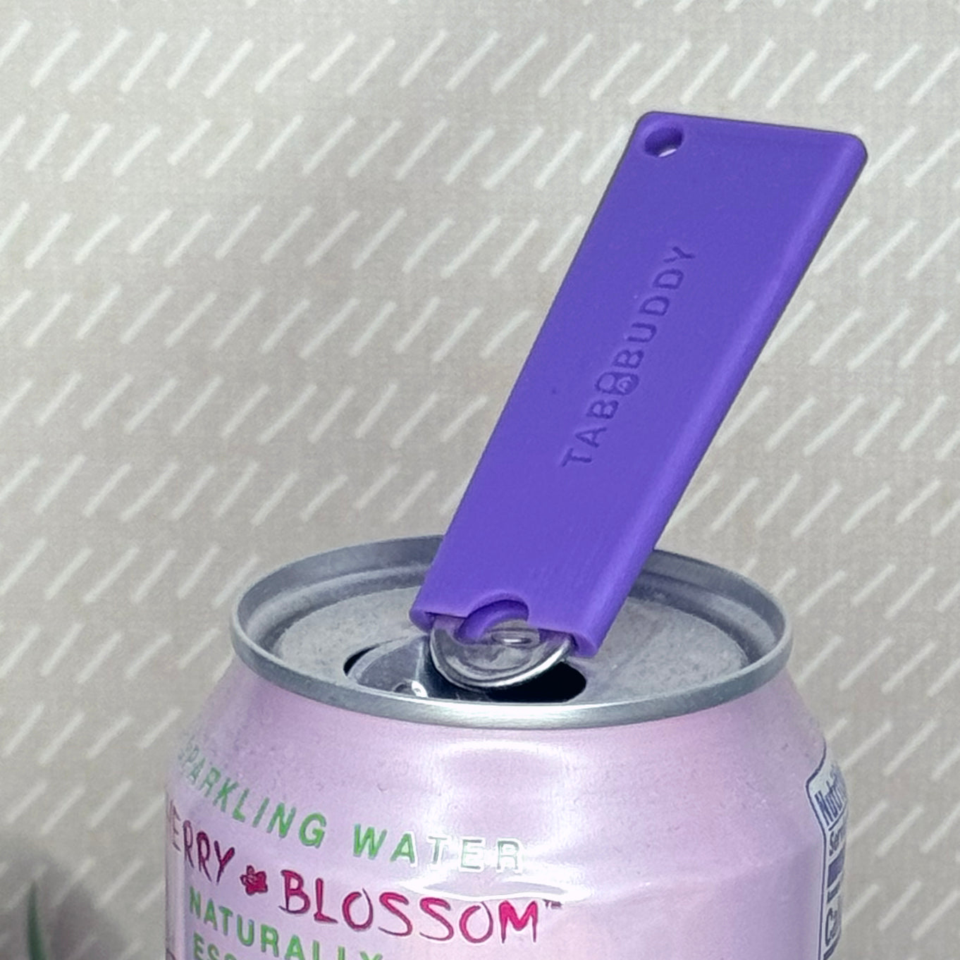 A purple Tab Buddy Classic soda can opener rests atop the tab of an open can of sparkling water with a beige background.