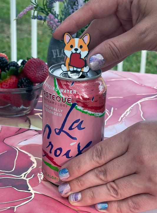 Person holding a soda can with a dog-shaped Tab Buddy on a pink surface.