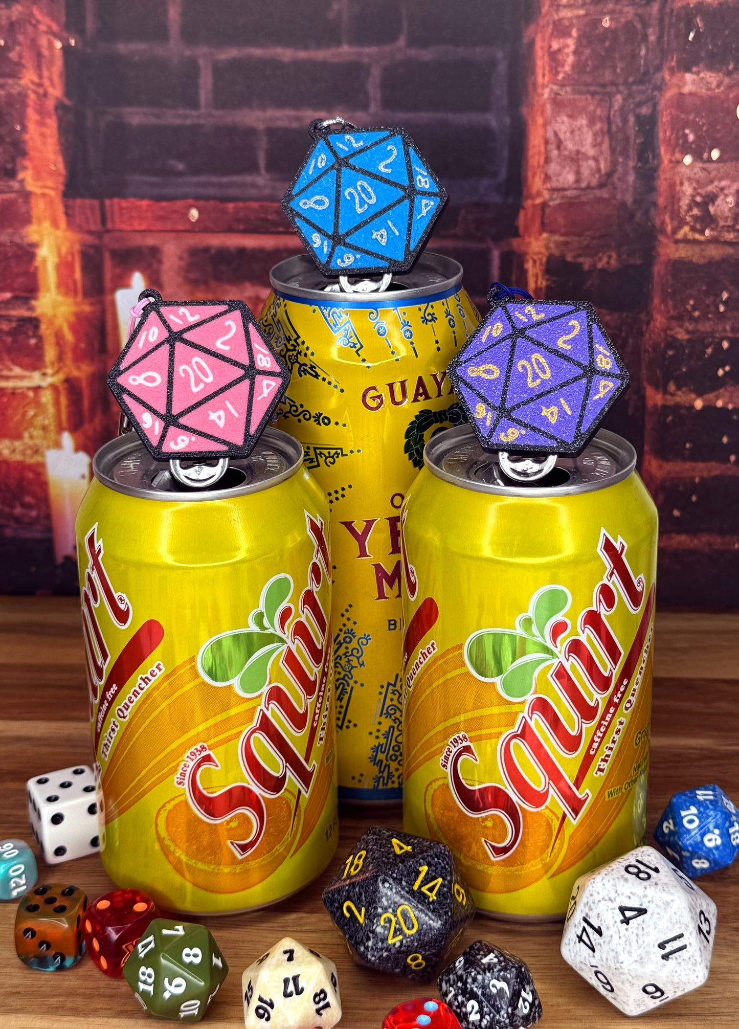 Three squirt soda cans with D20 dice Tab Buddies on top against a brick wall background