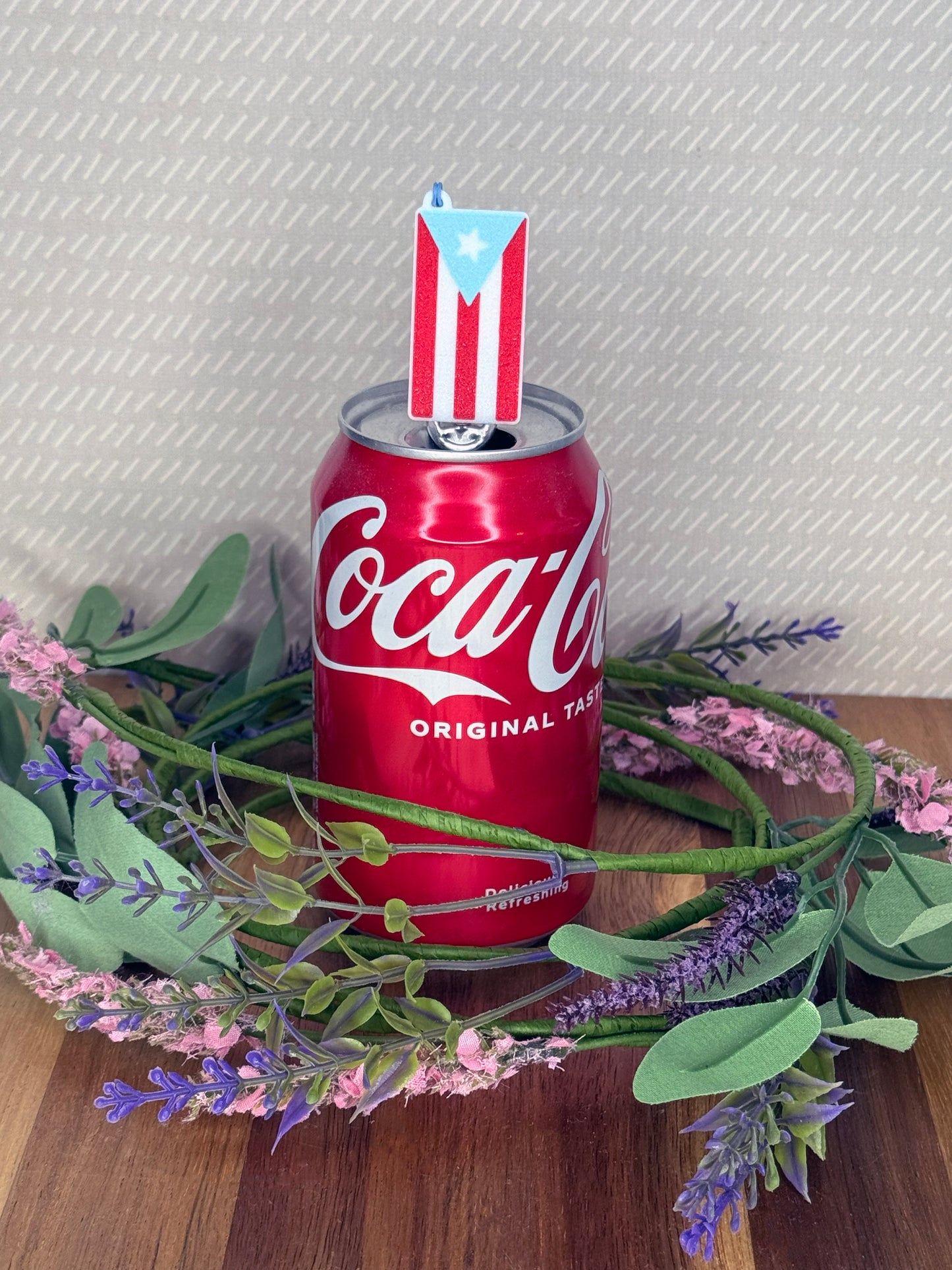 A red, white and light blue Pureto Rico flag Tab Buddy soda can opener rests atop the tab of an open can of sparkling water with a beige background and floral accents.