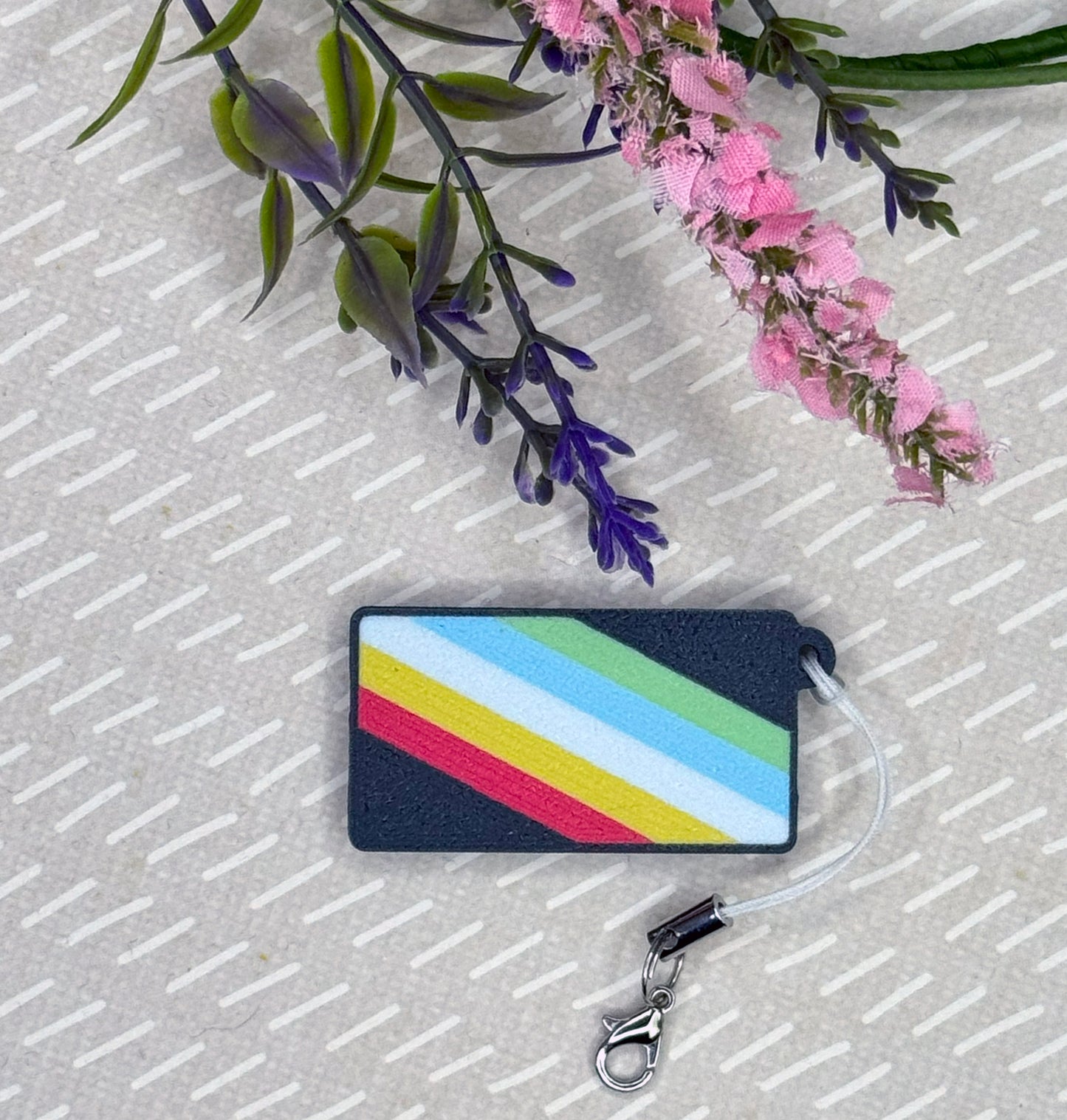 Tab Buddy with the disability pride flag on it with a strap attached rests on a beige surface with flowers.