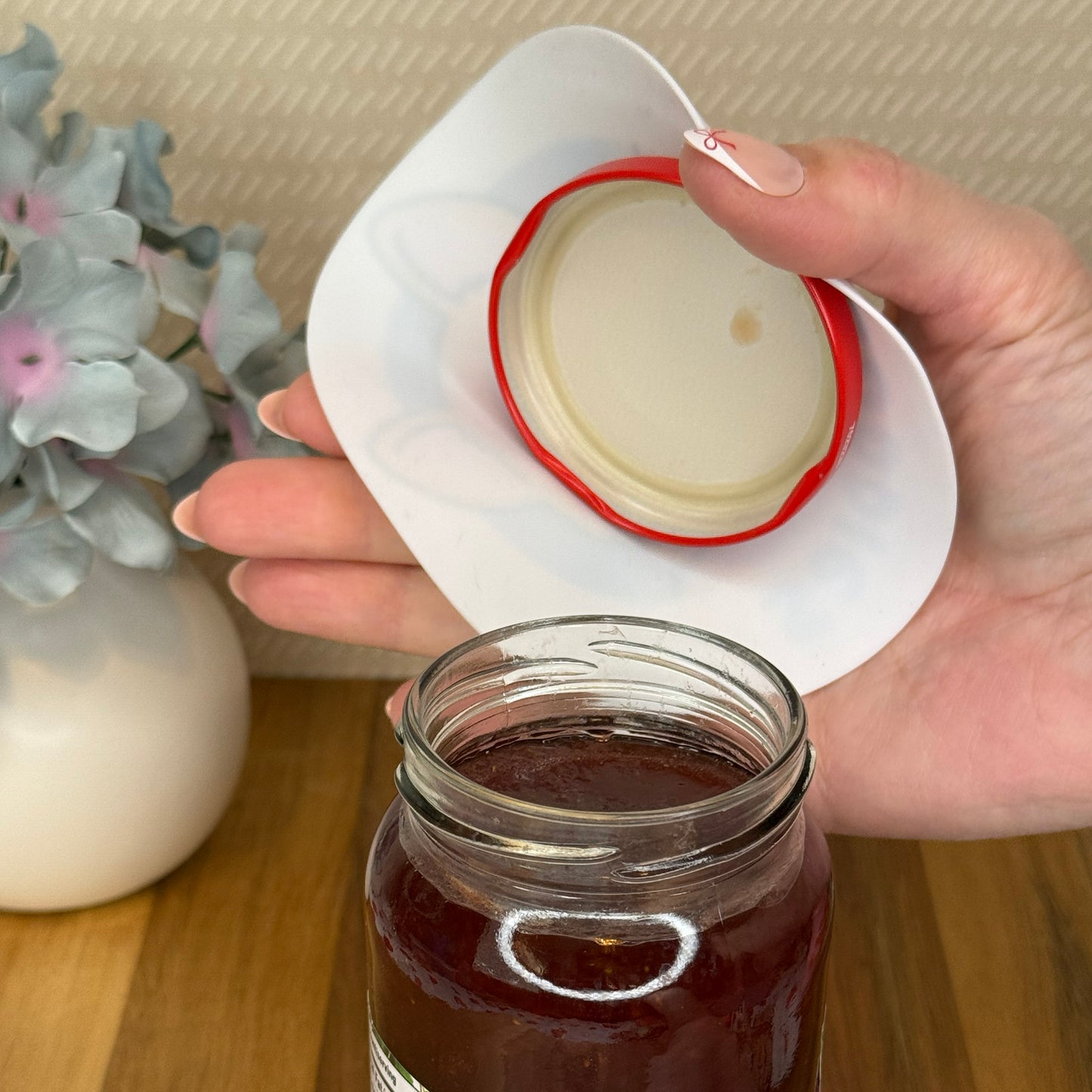 Hand holding Tab Buddies grippy pad and an open lid over a jar of strawberry preserves on a wooden surface.