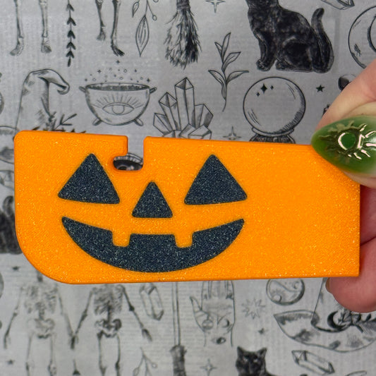 Orange Halloween-themed Tab Buddy XL with a black jack-o'-lantern face design, held by a hand against a patterned background.