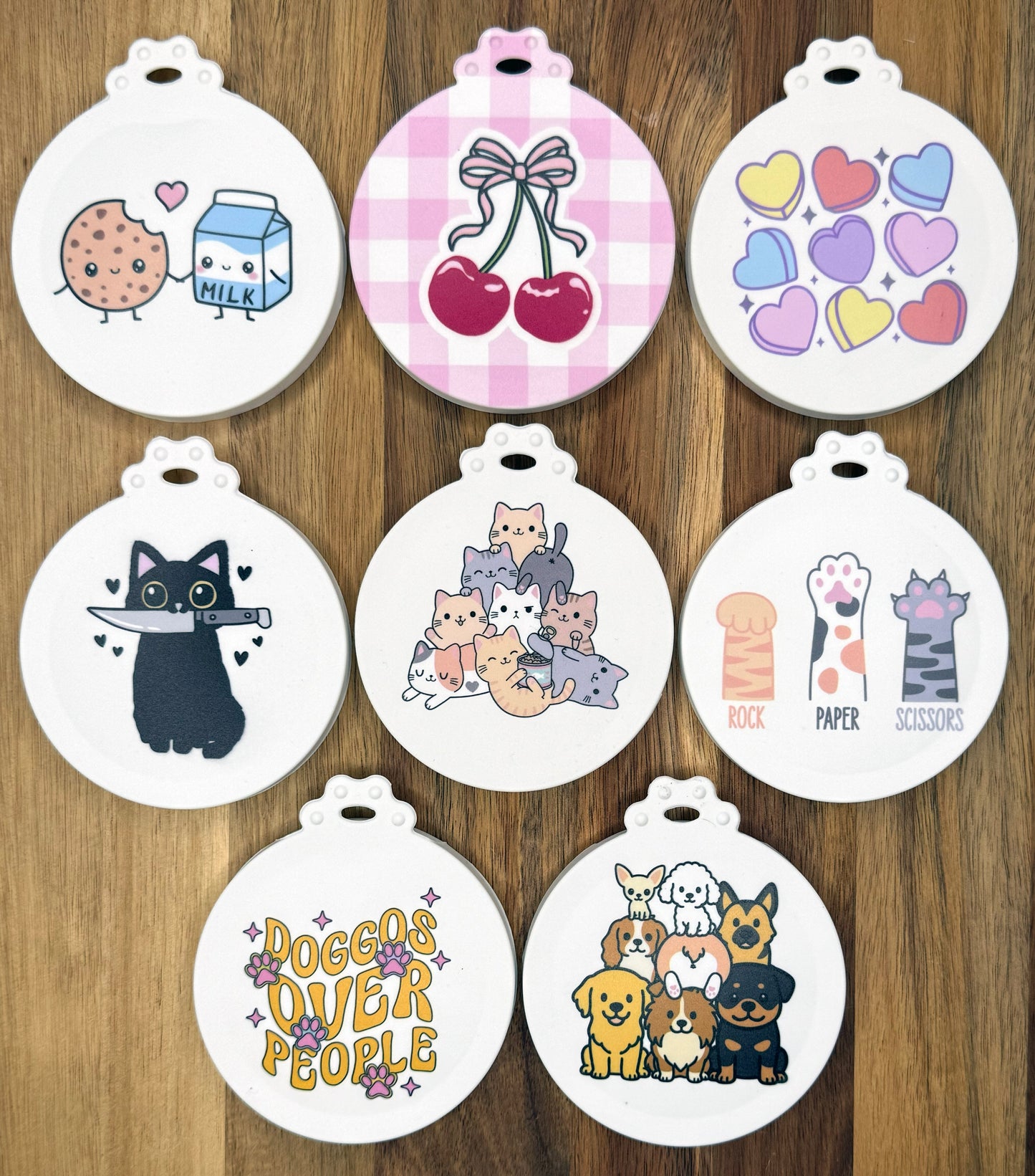 A group of 8 Tab Buddies reusable can lids available in Spring 2026 are shown on a wooden surface. Designs include themes of food, cats and dogs.