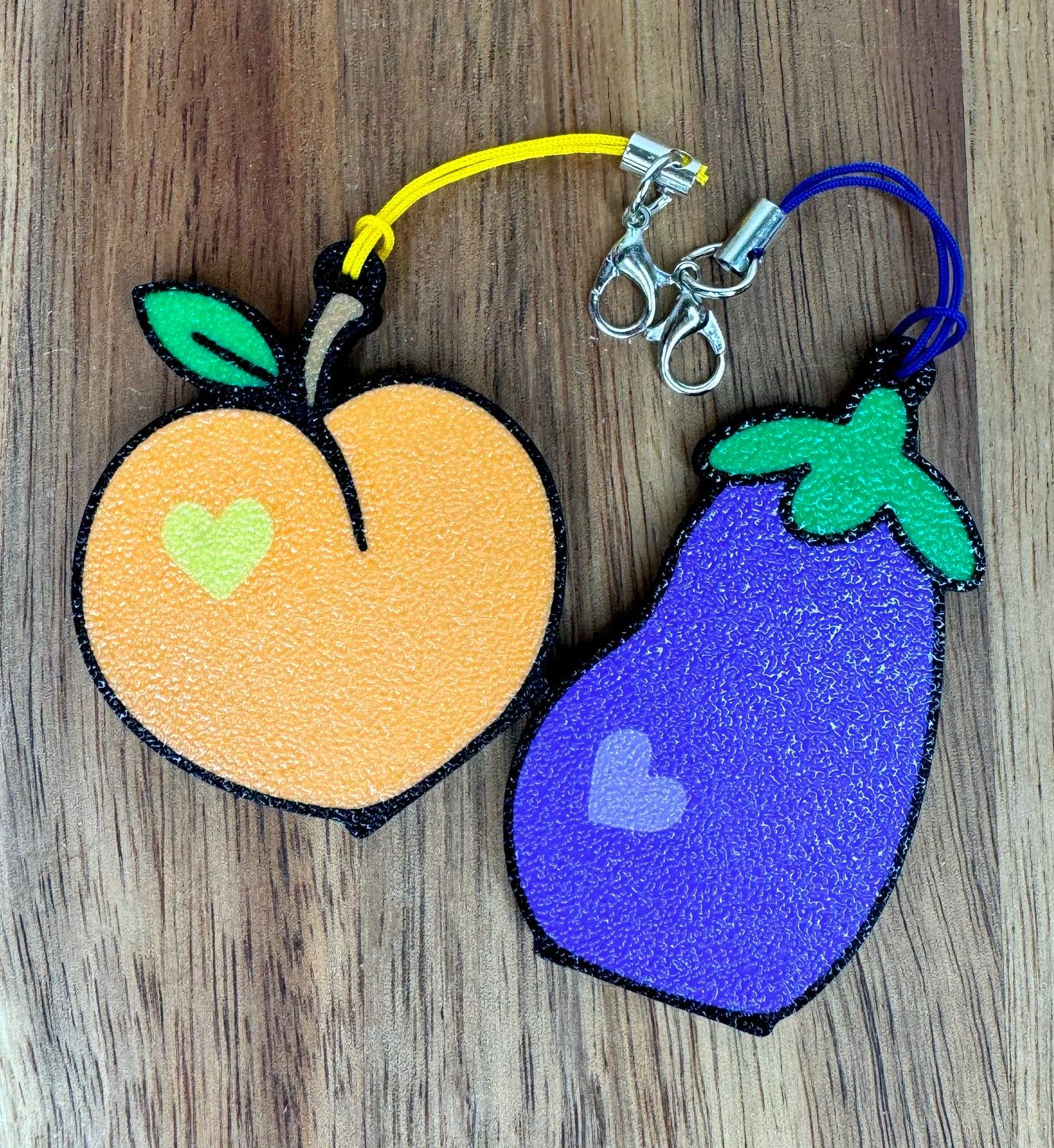 Two Tab Buddy keychains shaped like an peach and an eggplant on a wooden surface
