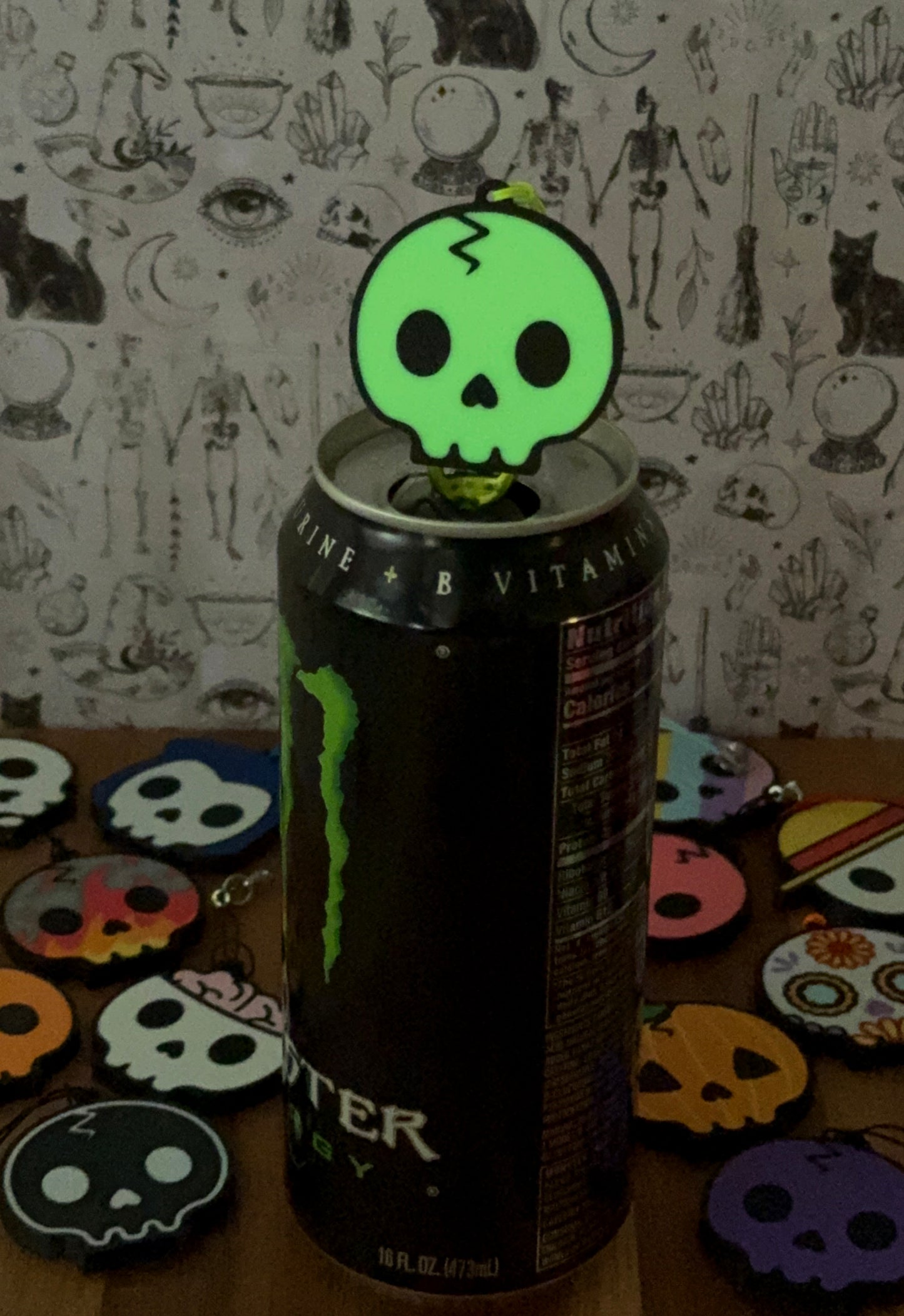 Monster energy drink can with a green skull-shaped Tab Buddy glowing on top. Other skull design Tab Buddies are in low-light background.