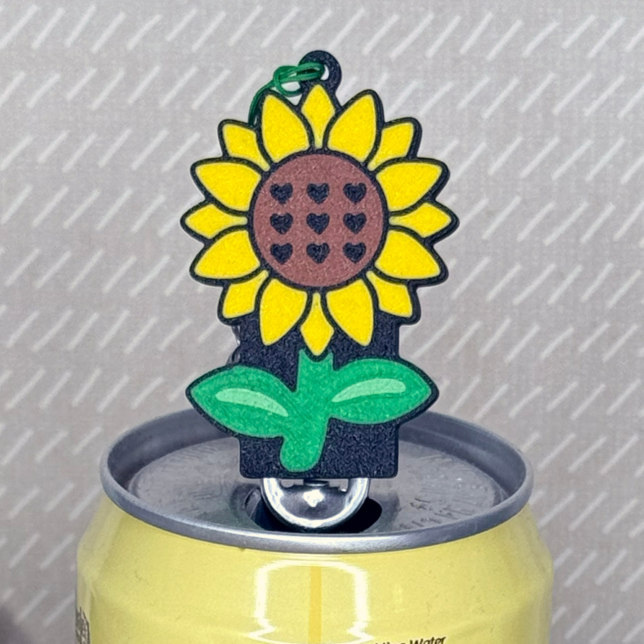 A yellow, brown and green sunflower-shaped Tab Buddy soda can opener rests atop the tab of an open can of sparkling water with a beige background.
