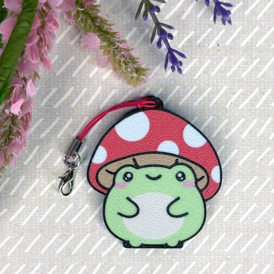 Cute green frog Tab Buddy with red polka dot mushroom on a textured surface with flowers.