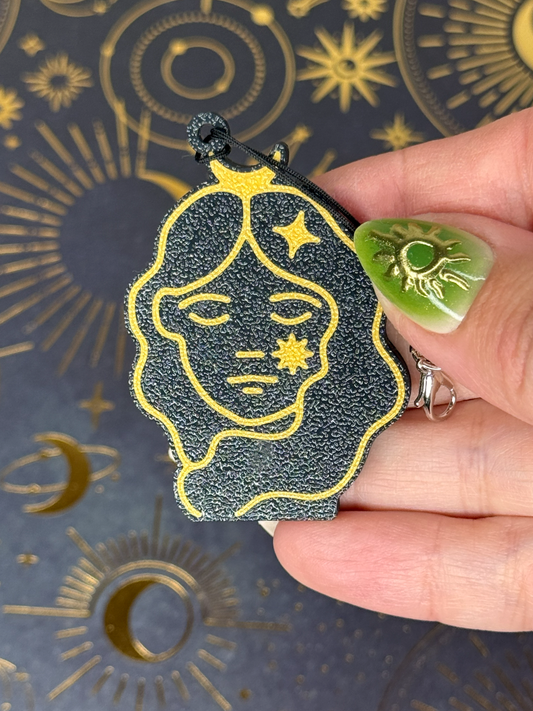 Hand holding a black and gold Virgo Zodiac face design Tab Buddy against a celestial pattern background.