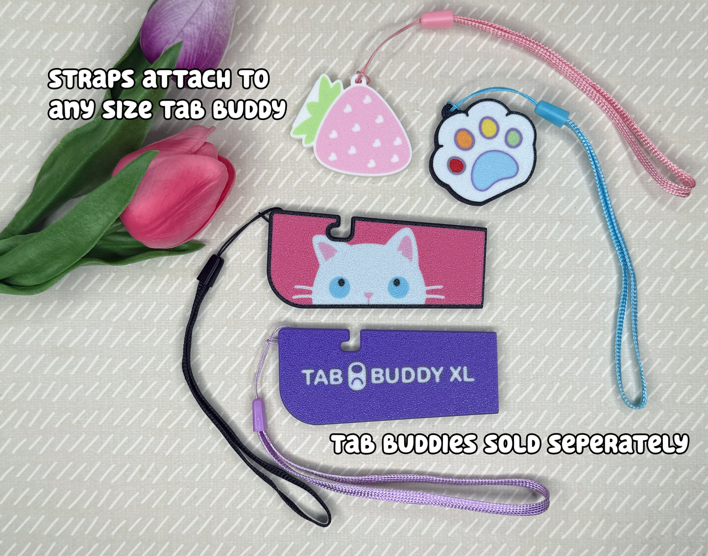 Four various Tab Buddies are shown with optional wrist straps attached. Text reads, "Straps attach to any size Tab Buddy. Tab Buddies sold seperately."