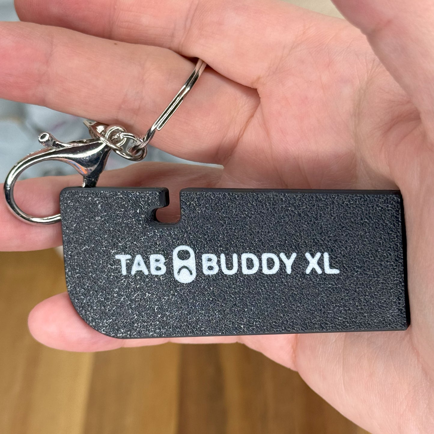 Black 'TAB BUDDY XL' is shown on a keychain held in a hand with a blurred background.