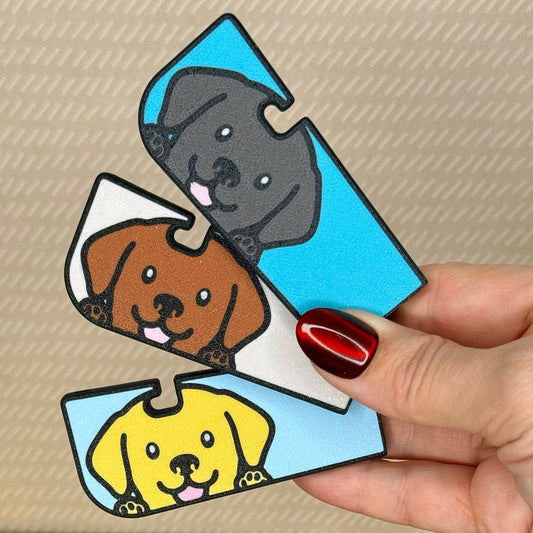 A hand holds three labrador-inspired Tab Buddy XLs in black, chocolate and yellow color variations in front of a tan background.