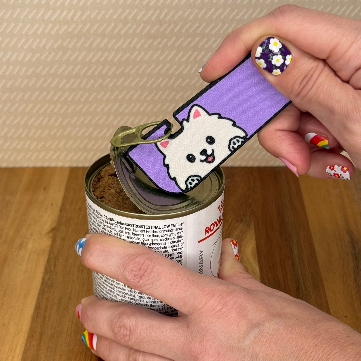 Hand using a Tab Buddy XL with a Pomeranian dog design to open a can of dog food on a wooden surface.