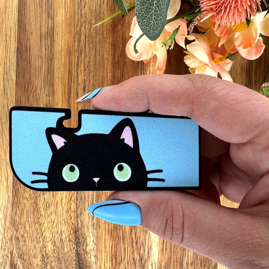 Hand holding a Tab Buddy XL with a black cat design on a wooden surface with flowers in the background