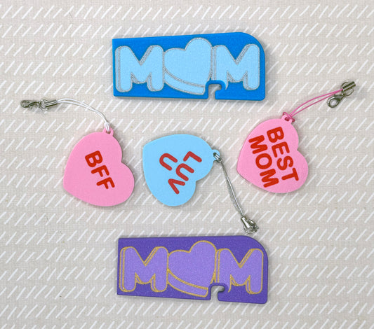 Various colorful heart and Mom-themed soda Tab Buddies and Tab Buddy XLs on a textured beige background