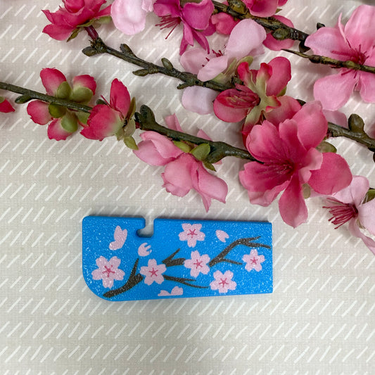 A Tab Buddy XL in Cherry Blossom design variation rests atop a beige surface with cherry blossom flowers in background.