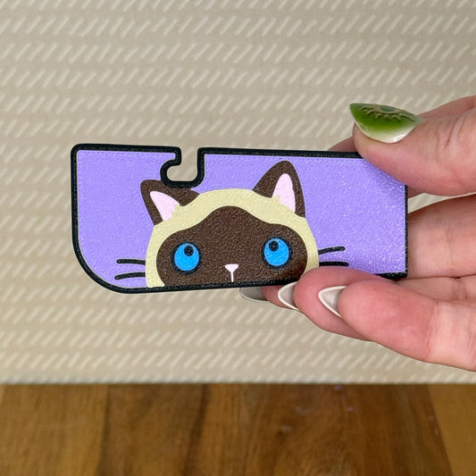 A sneaky cat Tab Buddy XL is held in a hand against a beige background. The cat is a blue-eyed siamese peeking up along the edge of the XL with a purple background.