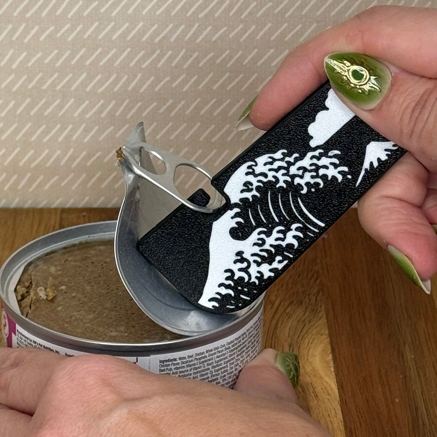A Tab Buddy XL with a black and white Great Wave design is used to peel back the tab lid on a can of cat food.