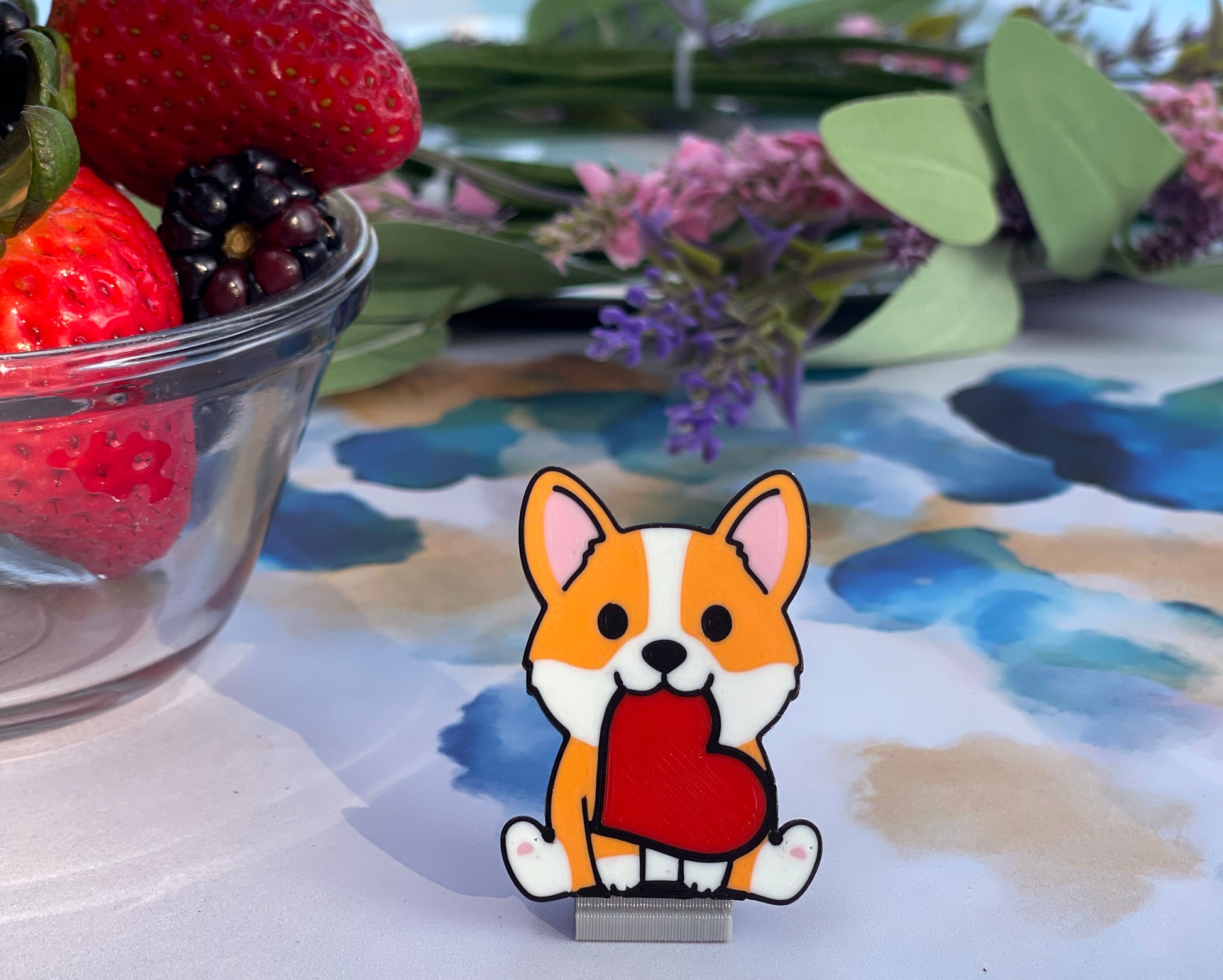 Cartoon corgi with a heart Tab Buddy in front of a bowl of strawberries and flowers on a floral-patterned tablecloth.