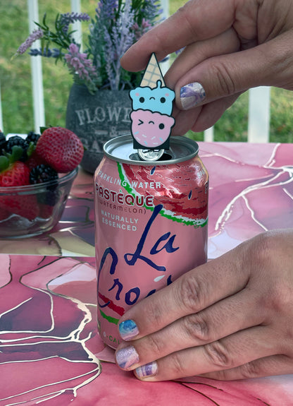 Person holding a pink soda can with a ice cream Tab Buddy on top, surrounded by strawberries and flowers on a pink surface.