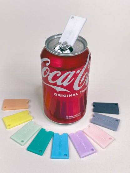 Coca-Cola can with a white Tab Buddy Mini on top and a collection of colorfulTab Buddy Minis surroundig the can on a light gray background