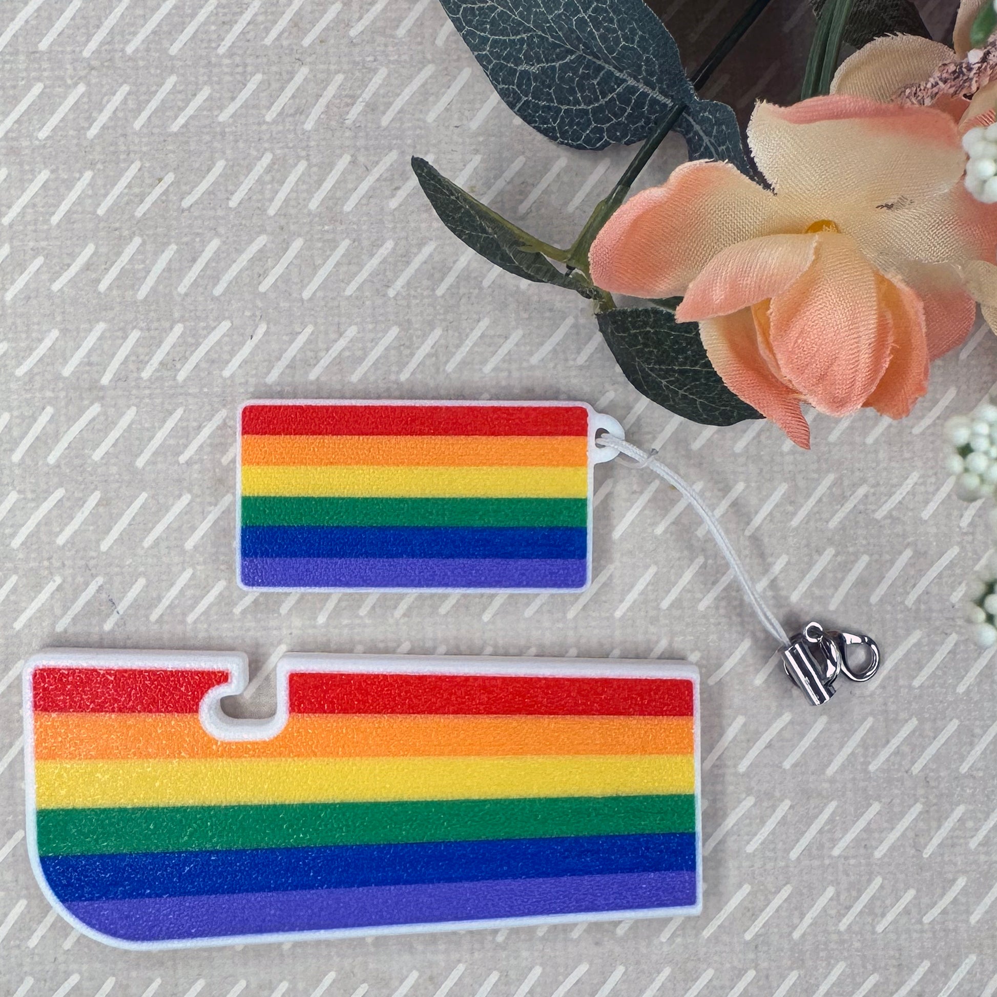 Two rainbow-colored Tab Buddies on a textured surface with flowers in the background.