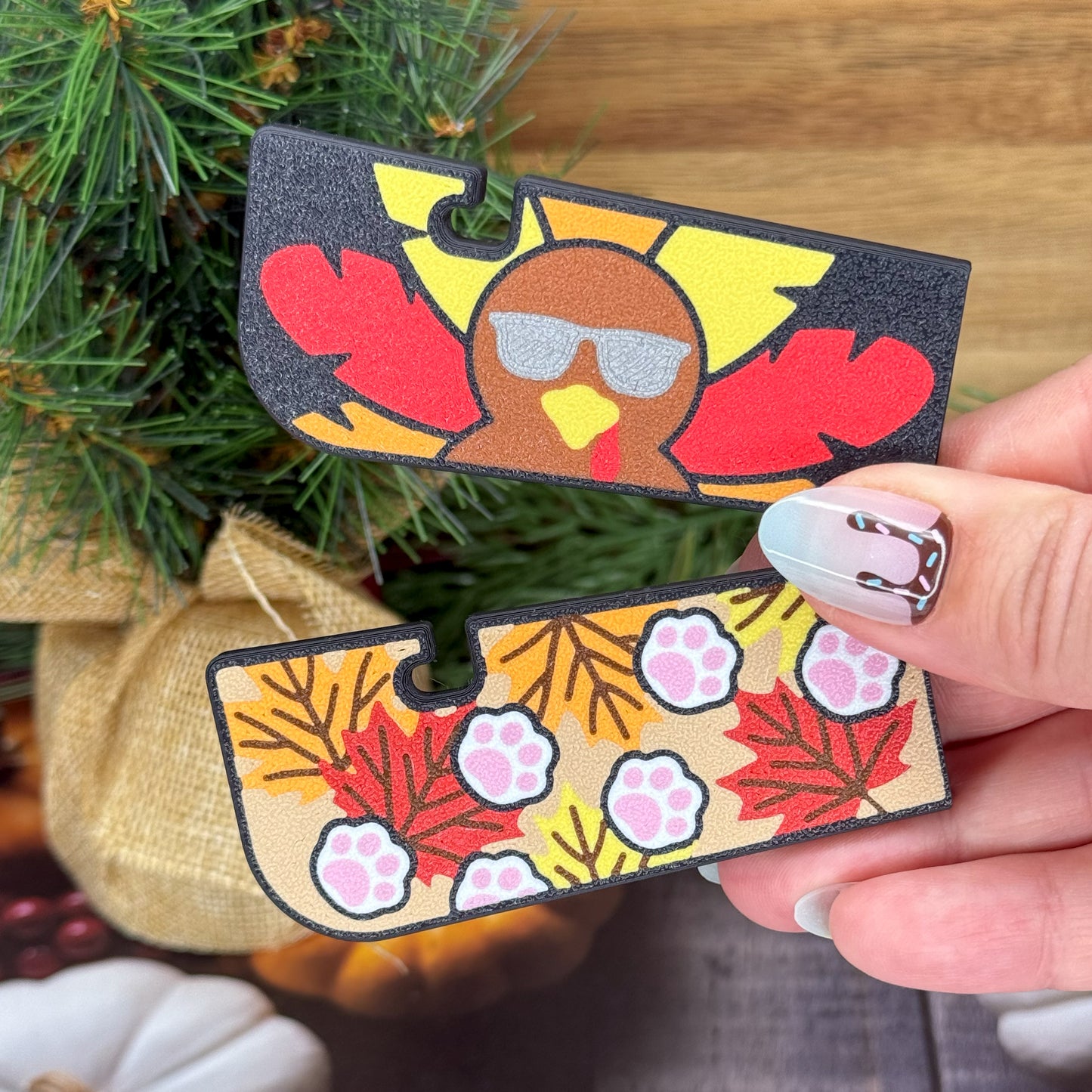 Hand holding two decorative Tab Buddies with turkey and aw print design sin front of a tree.