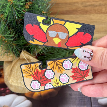 Hand holding two decorative Tab Buddies with turkey and aw print design sin front of a tree.