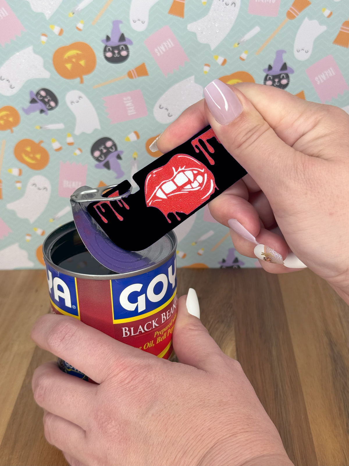 An individual with long nails uses a black Vampire Lips Tab Buddy XL to open a can of black beans.