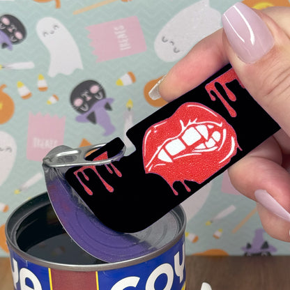 A hand with pale pink manicured nails uses a black Vampire Lips Tab Buddy XL to open a can of black beans.
