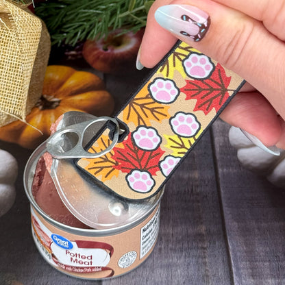 Hand holding a Tab Buddy XL with paw prints on a fall leaves design over a can of potted meat.