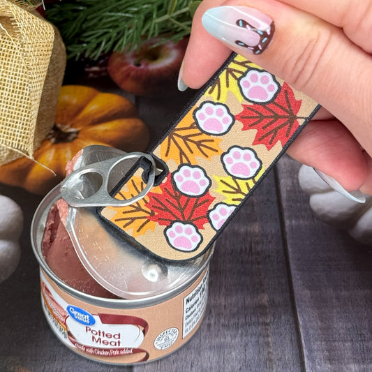 Hand holding a Tab Buddy XL with paw prints on a fall leaves design over a can of potted meat.