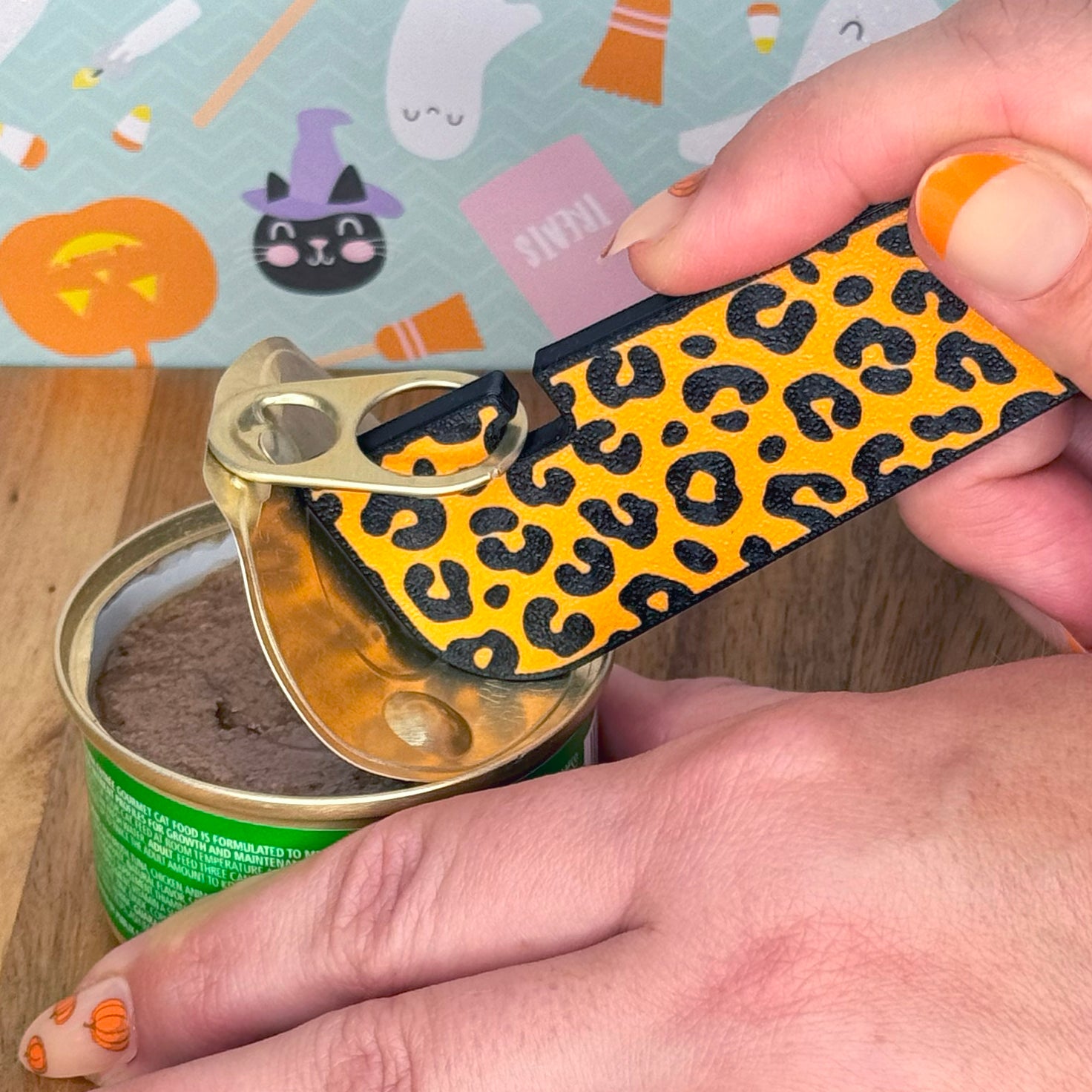 Hand using an orange leopard print Tab Buddy XL on a canned pet food with a Halloween-themed background.