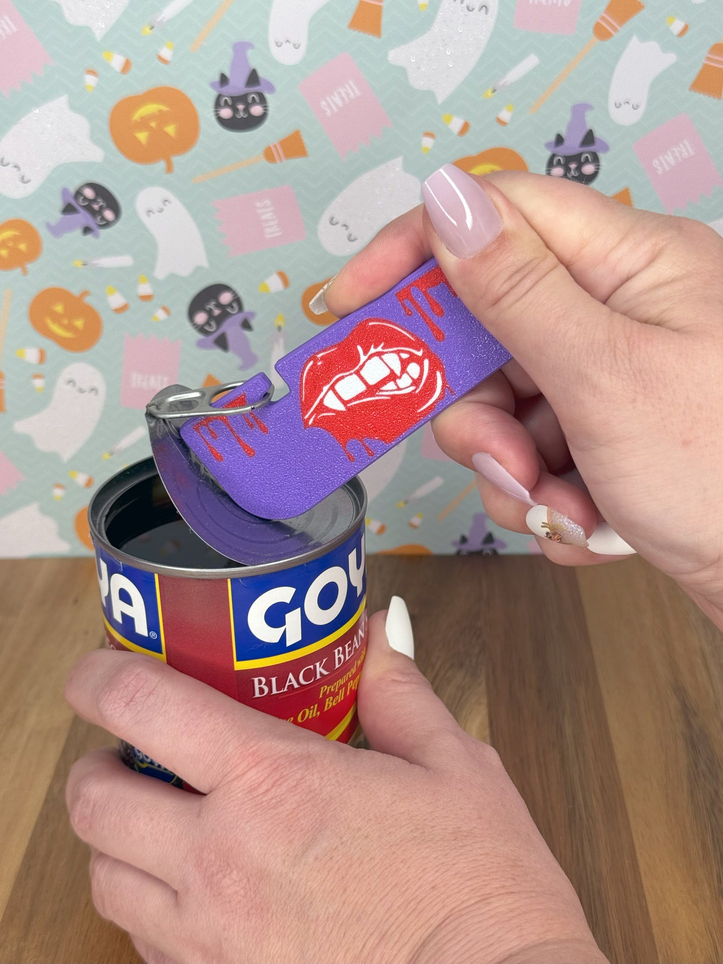 Hand holding Tab Buddy XL with a lips designs on it opening a can of Goya black beans with a Halloween-themed background