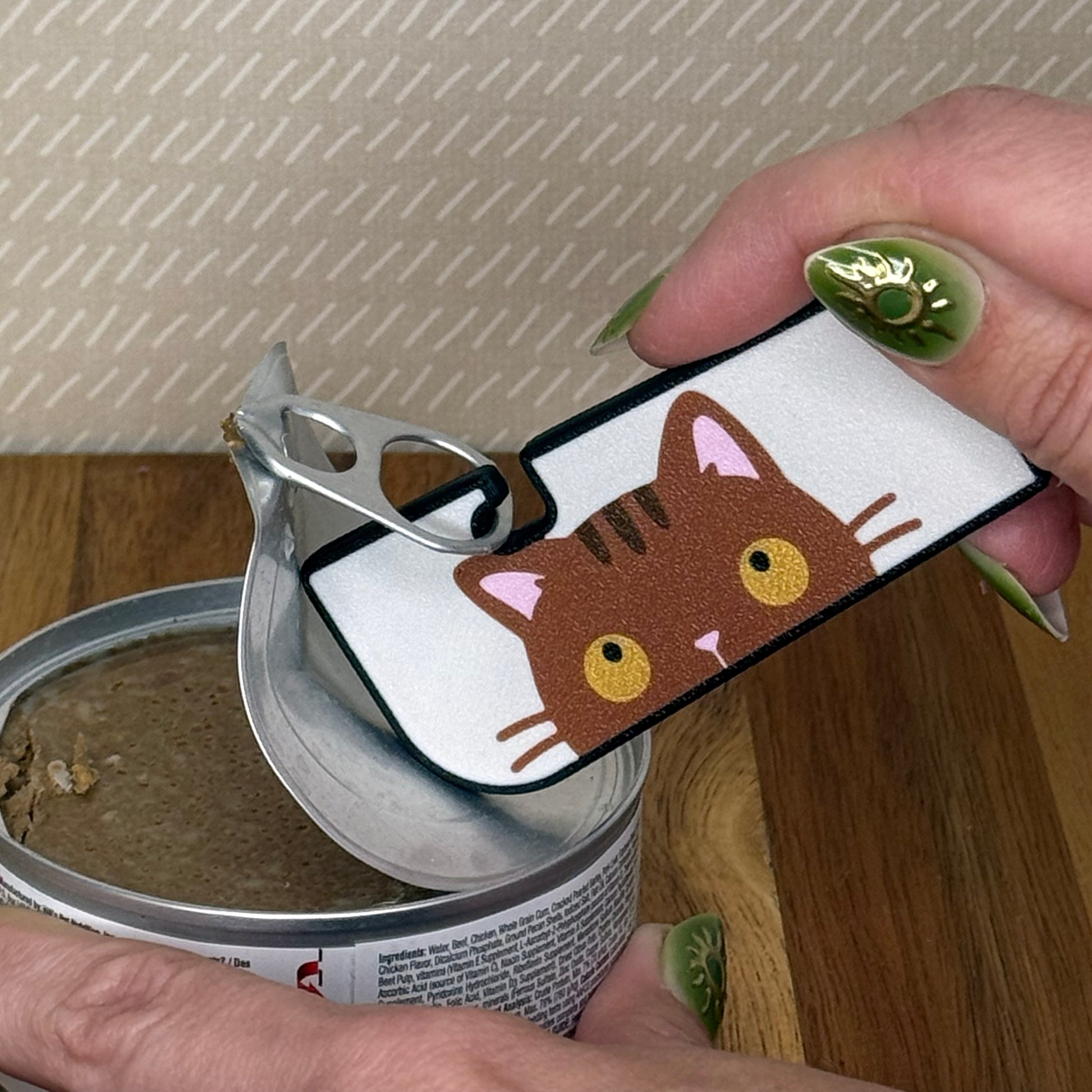 A Tab Buddy XL with a brown tabby cat design is used to peel back the tab lid on a can of cat food. 