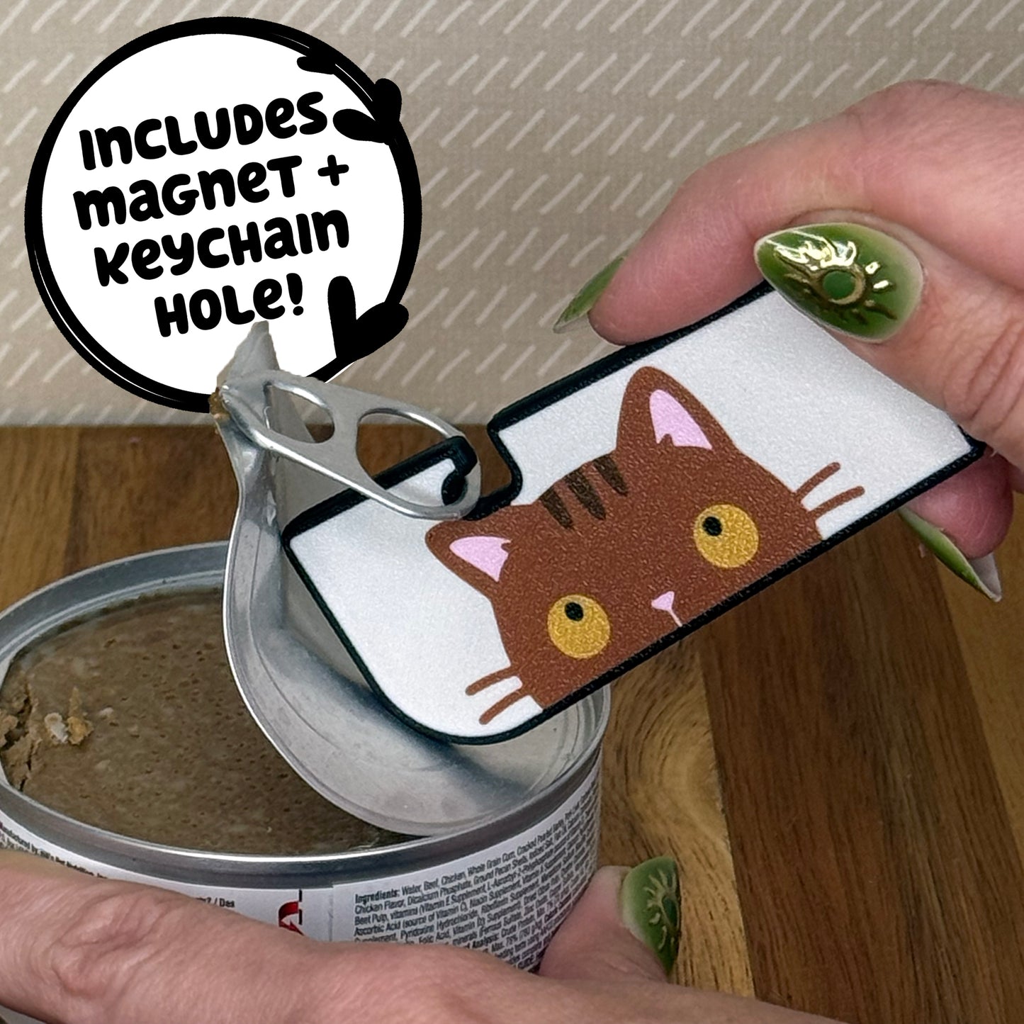 A Tab Buddy XL with a brown tabby cat design is used to peel back the tab lid on a can of cat food. Text reads "Includes magnet + keychain hole!"