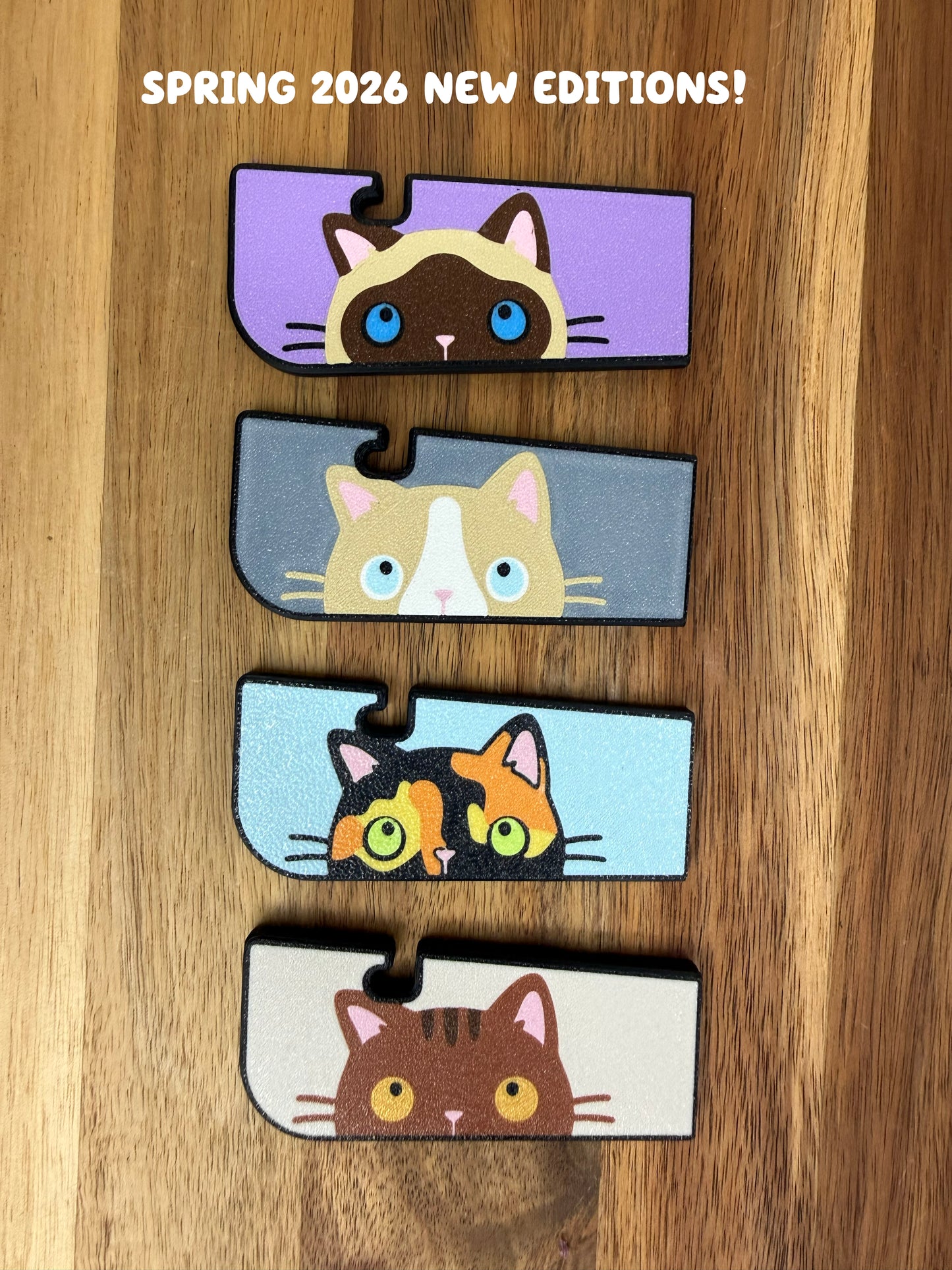 Four Tab Buddy XLs in siamese cat, ragdoll cat, tortoiseshell cat and brown tabby cat color variations rest atop a wooden surface. Text reads "Spring 2026 new editions!"