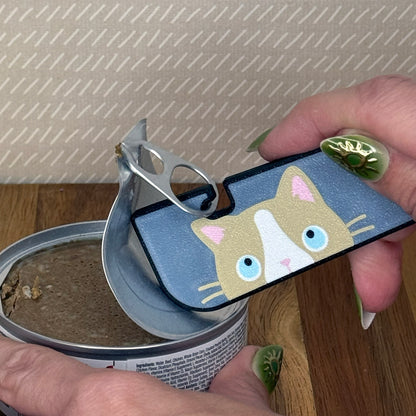 A Tab Buddy XL with a beige and white ragdoll cat design is used to peel back the tab lid on a can of cat food. 