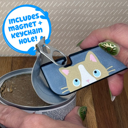 A Tab Buddy XL with a beige and white ragdoll cat design is used to peel back the tab lid on a can of cat food. Text reads "Includes magnet + keychain hole!"
