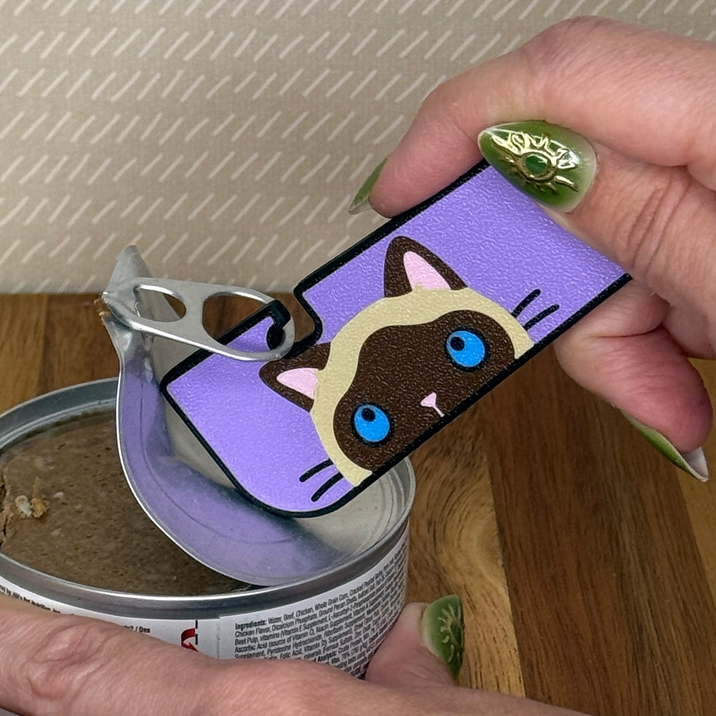 A Tab Buddy XL with a brown and tan siamese cat design is used to peel back the tab lid on a can of cat food.