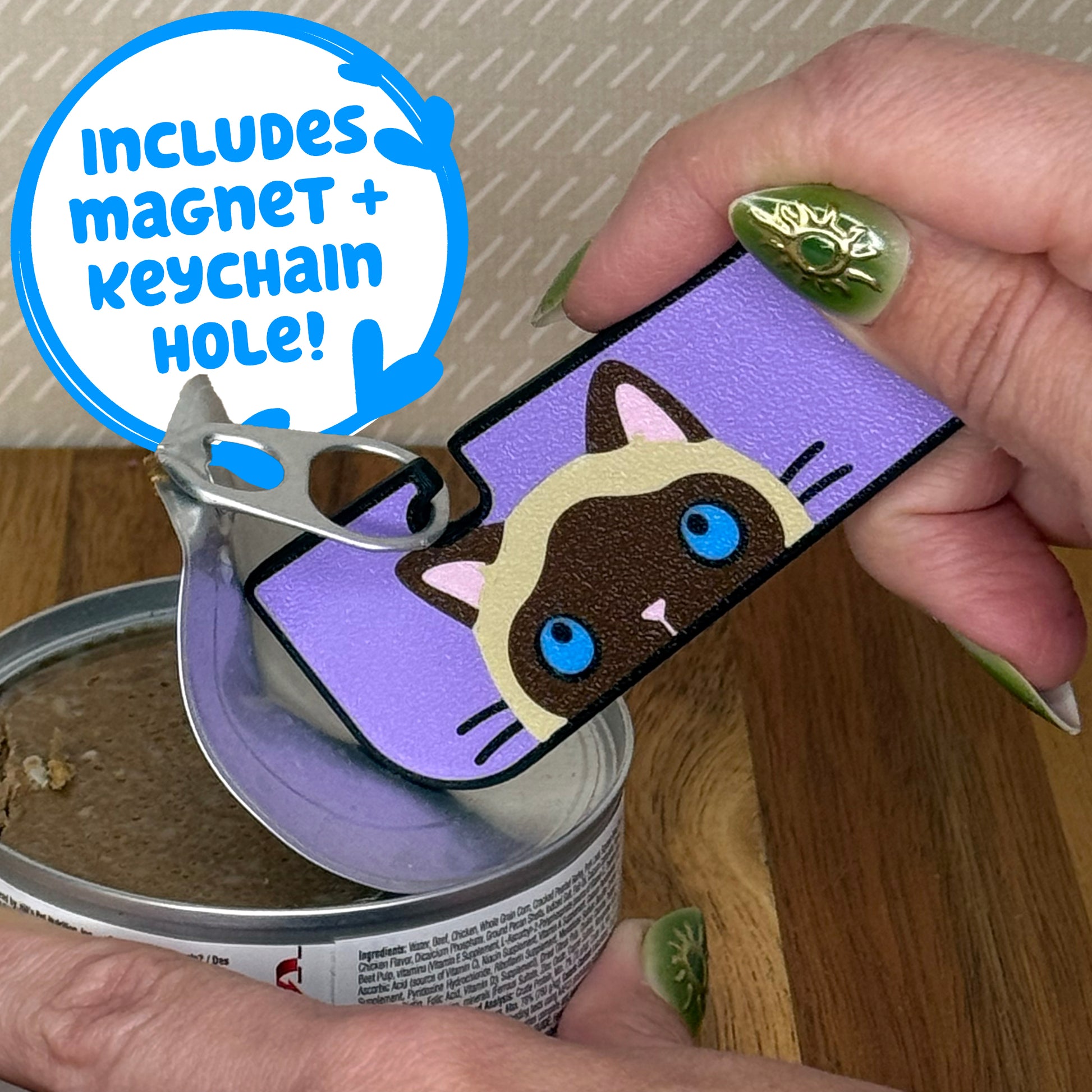 A Tab Buddy XL with a brown and tan siamese cat design is used to peel back the tab lid on a can of cat food. Text reads "Includes magnet + keychain hole!"