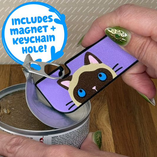 A Tab Buddy XL with a brown and tan siamese cat design is used to peel back the tab lid on a can of cat food. Text reads "Includes magnet + keychain hole!"