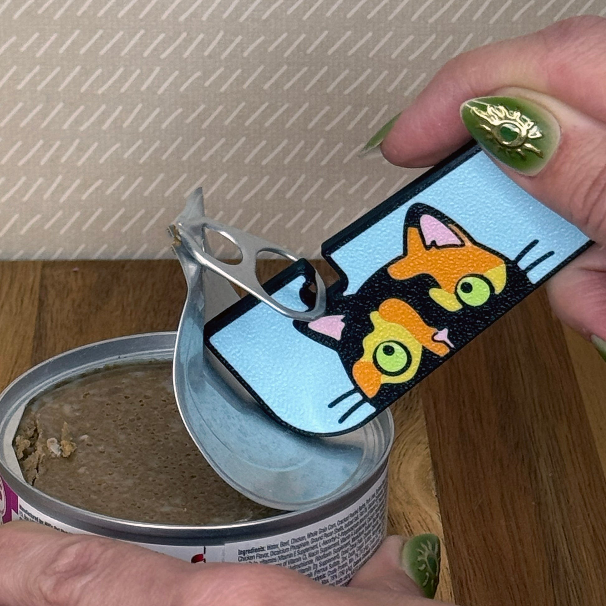 A Tab Buddy XL with a black, orange and yellow tortoiseshell cat design is used to peel back the tab lid on a can of cat food. 